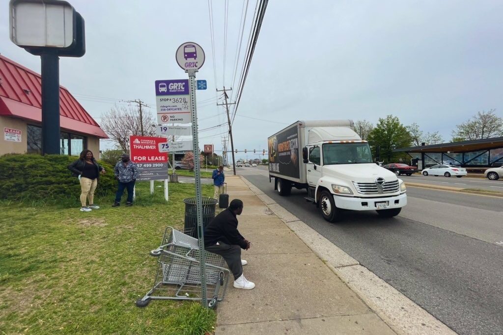 A man sitting on a shopping cart due to there being no basic transit infrastructure at a bus stop.
