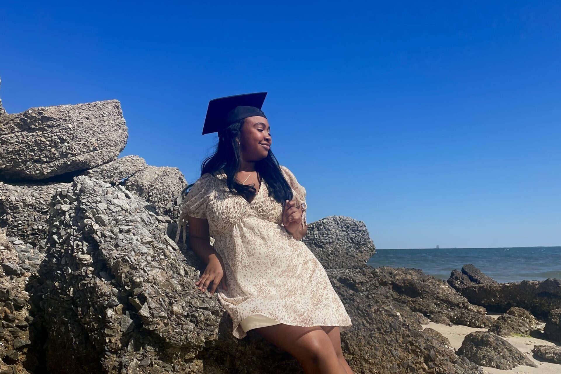 Victoria Jenkins taking a graduation photo on a beach, leaning up against several large rocks.
