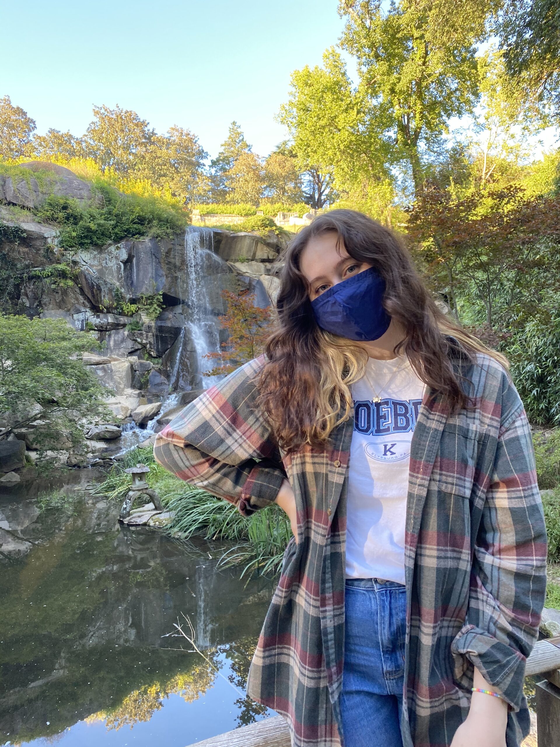 Katherine Hewes outside with a mask and wearing a flannel in front of a waterfall and mountains